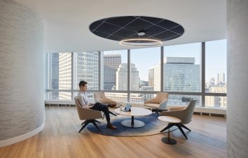 A man sits reading in a common area with views of downtown behind him