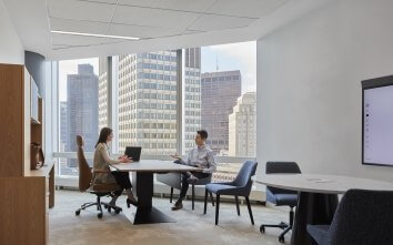 Two people chat in an office room with chairs, desks and a downtown view around them